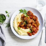 Een licht grijs bord staat op een witte tafel. Hierop is een schep smeuïge puree open geschept met daarbij de balletjes in tomatensaus. Het gerechtje is afgewerkt met wat verse peterselie. 