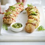 Twee broodjes liggen op een snijplank. Je ziet dat het twee versies zijn, nl een broodje met groene pesto, sneetjes ham en kaas. En dan nog een versie met roomkaas, paprika en rode ui. Extra pesto staat in een potje bij de broodjes, voor wie wat extra wil. Een vrouw zet de plateau of snijplank op tafel, haar handen zijn nog in beeld. 