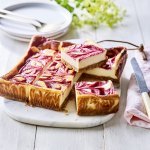 Een gebakken kaastaart staat op een marmere snijplank, het is wel een speciale cheesecake. Nl, de bovenzijde bevat frambozencoulis, waardoor er bovenaan een marmerpatroon is van de kaas en framboosjes. Echt een pareltje om naar te kijken. De taart is al aangesneden om onmiddelijk aan te vallen. 