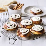 Op een taartrooster liggen 6 koekjes af te koelen. Het zijn koekjes van cinnamon rolls, een nieuwe versie om te genieten van deze heerlijke en kruidige zoetigheid. Op de koekjes is frosting gespoten op basis van roomkaas. 