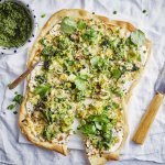 Op een witte snijplank ligt de rechthoekige flammkuchen. Hij heeft prachtige groene tinten vavn de erwtjes, prei en blaadjes waterkers. Er ligt een groot mes langs om aan te snijden een potje met extra pesto van waterkers.