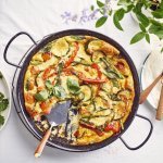 De frittata staat op tafel in zwarte pan, waardoor hij nog mooier oogt dan dat hij al is. De tomaatjes, paprika, courgette en asperges geven leuke kleurtjes aan de frittata. Er staat een slaatje langs de linkse zijde, met komkommer, radijsjes en verse kruiden.  