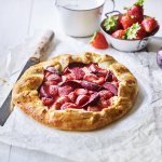 Een klaargemaakte galette staat op een houten witte tafel. Hij is prachtig goudgeel gebakken en het fruit oogt zoet-rode tinten. Er liggen nog wat aardbeien langs, alsook een paarse pruim.