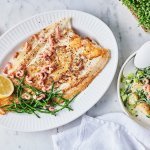 Een heerlijk krokant visje afgewerkt met zeekraal ligt op een groot bord. Er zijn enkele garnaaltjes aan toegevoegd die mooi roze blinken. De aardappelsalade staat er langs in een witte kom met witte lepel. Om af te werken ligt er 1 citroen doormidden gesneden.