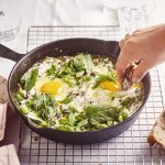 Op een rooster staat een zwarte gietijzeren pan met grieks getint gerechtje. Groene shakshuka met gebakken yoghurt. Er is een hand met een stukje brood in beeld dat er een dipje van zal nemen. 