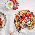 Een aangesneden taart staat op tafel met een wit laken er op. De kleuren van het rood fruit geven hierdoor nog een mooier zomers kleur. Naast de havermouttaart staat een bordje met blauwe spikkels en hier het stukje taart erop en een glaasje met de topping van de platte kaas en citroenzeste. 