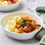 Op tafel staan twee uitgeschepte porties van het hoofdgerecht. Een schep heerlijke puree met kalfsfricassee. In de fricassee zitten spekblokjes, selder en wortelstukjes. Het gerechtje is afgewerkt met verse peterselie. Er ligt een licht groene servette langs en wit bestek.