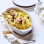 Op tafel staat een witte ovale ovenschotel met hierin de gele currysaus met stukjes appel en ananas. Hier bovenop liggen de bouten te blinken. De ovenschotel staat op een beige linnen handdoek met rijst, borden en een witte pepermolen langs.