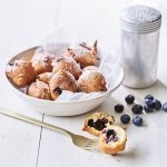 In een diep wit bord ligt een wit boterpapiertje met hierin enkele oliebollen. Er is eentje uitgeprikt en ligt op tafel doormidden gesneden, hier zie je duidelijk de blauwe bessen in zitten. Er is bloemsuiker over de oliebollen gestrooid voor een extra zoete toets. 