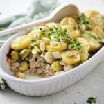 Een ovenschotel staat op het midden van de tafel, er is al een portie uitgeschept. Je ziet het gehakt onderaan liggen, met spruitjes , stukjes knolselder en goudgele aardappelschijfjes. 