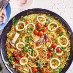 Een grote pan gevuld met een kleurrijke paella staat op het aanrecht. Hij zit vol met de goudgele rijst, in ringen gesneden pijlinktvis, kerstomaten, paprika en verse peterselie.  