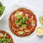 Op tafel staan twee klaargemaakte pizza's op een wit bakpapiertje. Hij is afgewerkt met verse rucola en wat olijfolie.