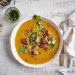 Op tafel staat een wit soepbord gevuld met heerlijk oranje pompoensoep. De soep is afgewerkt met verse groene kruiden, pompoenpitten en croutons van zuurdesembrood. Er ligt een witte linnen servette langs, geknoopt, en een soeplepel. 