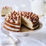 Op tafel staat een prachtige taart, het is een twist op de klassieke tiramisu. Hij staat op een witte taartschaal, en is afgewerkt met toefjes mascarpone en cacaopoeder. In de achtergrond staat een koffiekan en tasjes koffie.