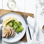 Op tafel ligt een houten snijplank met hierop een lichtblauw groot plat bord. Hierop is het hoofdgerecht geserveerd, stukjes varkenshaasje in sneetjes, plakjes broccoli en een een schep aardappelpuree. Het sausje zit in een sauskannetje langsheen het bord.