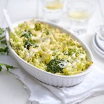 Een grote ovenschaal staat op een houte witte tafel. Hij staat op een wit lapje stof. De ovenschotel is gevuld met macaroni, broccolie en witte bloemkoolsaus. Er staan enkele bordjes langs met wit bestek om direct aan te vallen. 