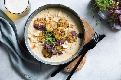 Balletjes van champignons en bonen met mosterdsaus in een diep grijs bordje op een ruwe houten plankje. Linksboven staat een versgetapt bier en rechts nog een portie extra groenten. Onderaan ligt nog een grijze linnen servette en zwart bestek langs het bord. 