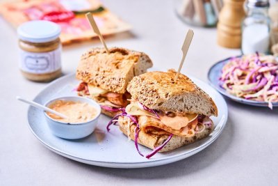 Er liggen twee broodjes op een licht blauwgrijs bordje. De broodjes zijn belegd met breydelspek, koolsla van rode kool en spitskool, een sneetje kaas en homemade saus. 