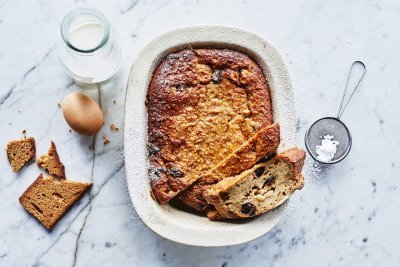 Een heerlijke homemade broodpudding staat op een marmer aanrecht in een witte ovenschaal. Hij is al aangesneden en een stukje uit de vorm genomen. Om de foto meer boddy te geven ligt er een eitje, flesje melk, stukje peperkoek en bloemsuiker naast.