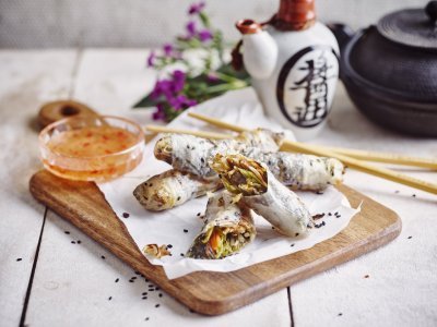 Heerlijke krokante springrolls liggen op een houten snijplank. Ze zijn vergezeld met een zoetpikante dipsaus. In de achtergrond staat nog een theepot en paarse bloemetjes.  