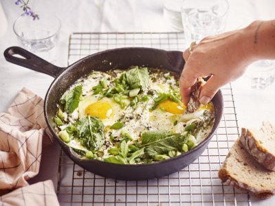 Op een rooster staat een zwarte gietijzeren pan met grieks getint gerechtje. Groene shakshuka met gebakken yoghurt. Er is een hand met een stukje brood in beeld dat er een dipje van zal nemen. 