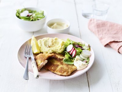 Een homemade shnitzel ligt op een pastelroze bord, samen met aardappelpuree, verse sla en zelfgemaakte mayonaise. Er is nog wat extra salade in een wit potje en mayonaise voor extra lekkers op te scheppen. 