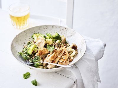 Een rijkgevulde quinoasalade boordevol verse groenten en kruiden zoals broccoli, paddenstoelen, rode ui, peterselie, koriander en munt. Bovenop de quinoa werd het varkensvlees geplaatst in schijfjes. Er staat een witbier bovenaan het bord, op de tafel.