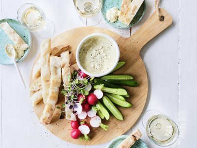 Op tafel staat een ronde houten snijplank met hierop een wit potje gevuld met onze pikante dip van yoghurt en witte bonen. Je kan de yoghurtdip gebruiken met stukjes snackkomkommer, brood en radijsjes in ons geval. Rond de plank staan enkele glazen witte wijn en helderblauwe broodbordjes. 