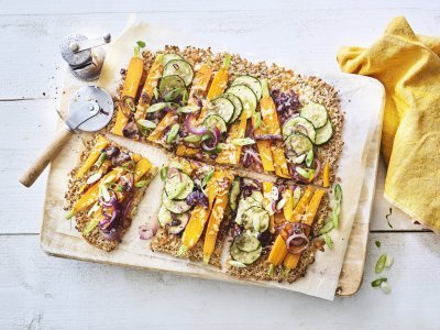 Een krokante bodem van bloemkool en walnoten waarop kleurrijke groentjes liggen zoals wortel, courgetteplakjes en rode ui. De plaattaart ligt op een houten snijplank waar een pizzasnijder langs ligt, de pepermolen en goudgele servette. 