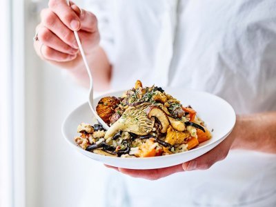 Vrouw staat te staren uit het raam met bordje risotto in haar hand. De risotto is afgewerkt met gebakken boschampignons en Brugge Oud.