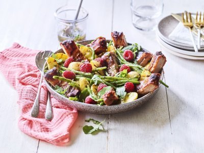 Een grote serveerschaal is gevuld met de aardappelsalade. Je ziet de framboosjes, boontjes en ribbetjes in de salade zitten. Er staat een torentje bordjes klaar met bestek om de salade te verdelen en op te eten. 