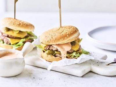 Twee heerlijke burgers liggen klaar op een marmere snijplank. Het vlees is verdeeld in twee dunne porties met de saus en gesmolten kaas er bovenop. De burgers worden recht gehouden door middel van een prikker.