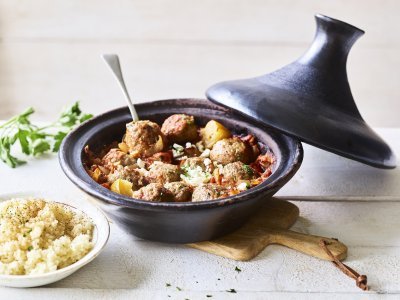 Zwartkleurige tajine met gehaktballetjes van lamsvlees, afgewerkt met verbrokkelde feta. Er staat een portie couscous naast de tajine. 