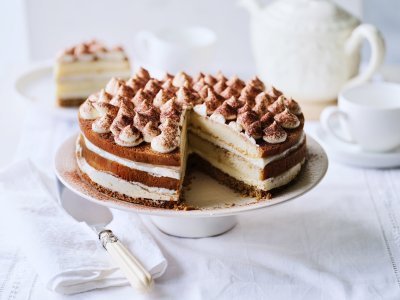 Op tafel staat een prachtige taart, het is een twist op de klassieke tiramisu. Hij staat op een witte taartschaal, en is afgewerkt met toefjes mascarpone en cacaopoeder. In de achtergrond staat een koffiekan en tasjes koffie.