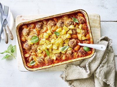 Op tafel staat een grote rechthoekige ovenschaal op een houten plank. Er staan witte bordjes langs met bestek om op te scheppen met een witte lepel. Bij de ovenschotel ligt ook een bruin servette. Het gerecht is afgewerkt met verse basilicum die mooi contrast geven op de tomatensaus en goudgele krokante aardappeltjes. 