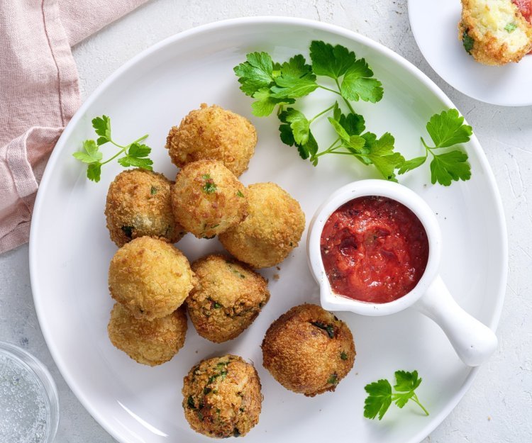 Heerlijke krokante balletjes liggen op een wit bord gepresenteerd. Hier is een klein potje met ketchup bijgeplaatst en enkele blaadjes peterselie voor extra groen. 