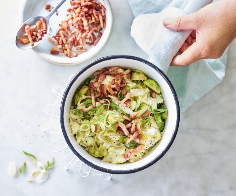 Smeuïge aardappelpuree met grofgehakte spruitjes en prei klaargemaakt in een kookpot. De kok in kwestie voegt de versneden lente-uitjes en gebakken spekjes toe als afwerking.