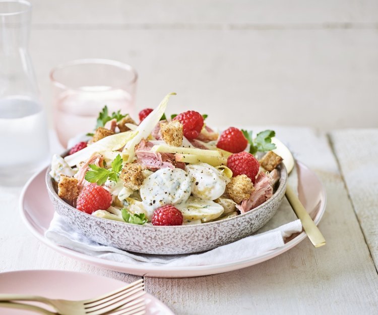 Een rijkelijke aardappelsalade is geserveerd in een grote kom. Je ziet de aardappel met de dressing, stukjes Breydelham, framboosjes en appel. De kom staat op een pastelkleurig roos bord. Er staat ook een karaf water langs met een glas in dezelfde tint als het roze bord. 