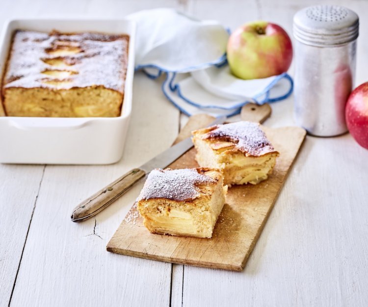 Een verse appelcake uit de oven, je reukt hem waarschijnlijk al. Er is een stukje uitgesneden en deze ligt opnieuw aangesneden op een houten plankje. Op tafel ligt een witte servette met blauwe randjes, nog enkele verse appels en de strooipot met bloemsuiker. 