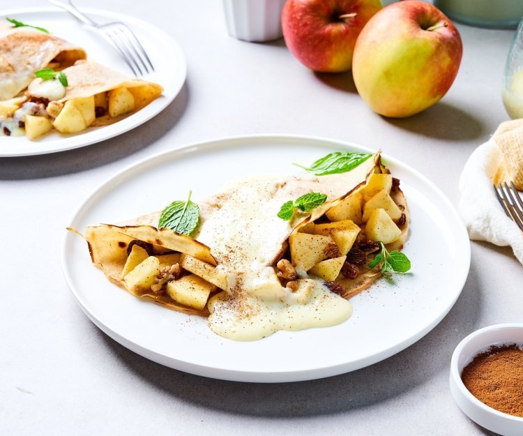 Op tafel staan twee borden met elk hierop een dichtgevouwen pannenkoek, die gevuld is met stukjes appel en rozijnen. Wij serveren hem met vanillesaus en verse blaadjes munt. Er liggen ook nog enkele appelen in de achtergrond en een restje vanillesaus.