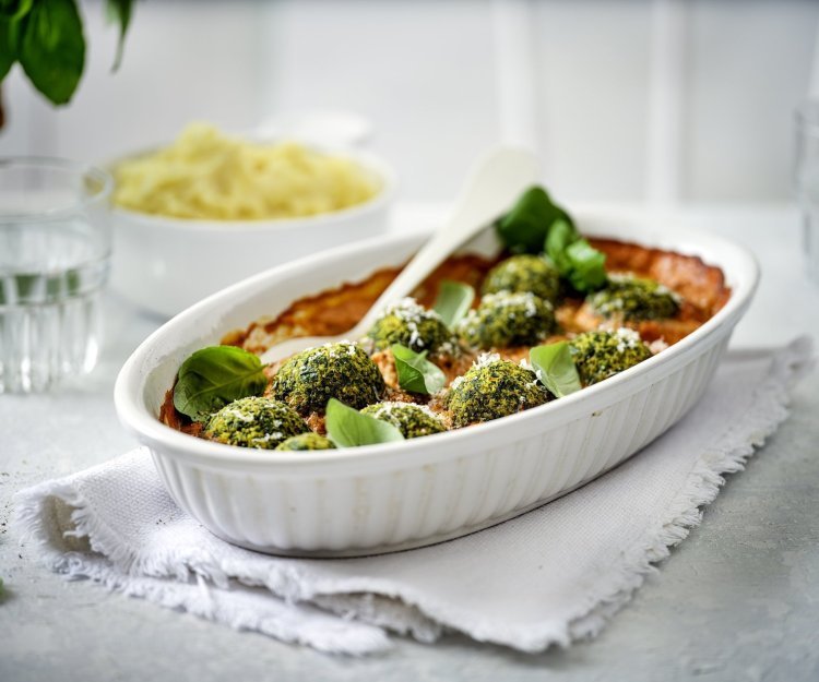 Een witte ovenschotel staat op tafel op een dikkere keukenhanddoek. De ovenschotel is gevuld met rode paprikasaus en hierop liggen de groene vegetarische balletjes van boerenkool. In de verte zie je ook nog een kommetje puree, klaar om aan tafel te gaan dus!