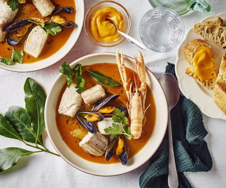 Een heerlijk kleurrijk bordje bouillabaisse staat op tafel. Wij maakten hem met topproducten van hier. Er ligt een donker groene linnen servette langs en bordje stukjes brood en verse rouille.