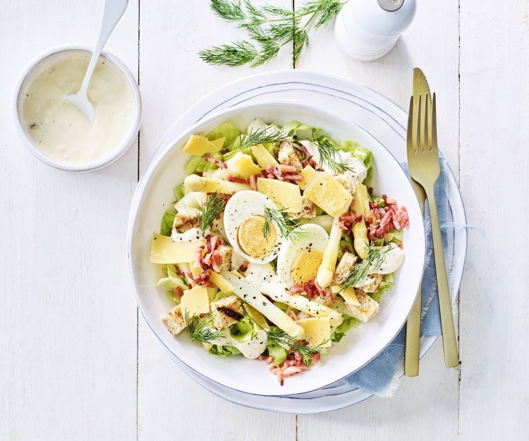 Een rijkgevulde salade staat klaar op tafel. Het witte bord is gevuld met sla, gekookte eitjes, stukjes asperges en kaasschilfers. Dat is de lente proeven in je bord, als je het mij vraagt!