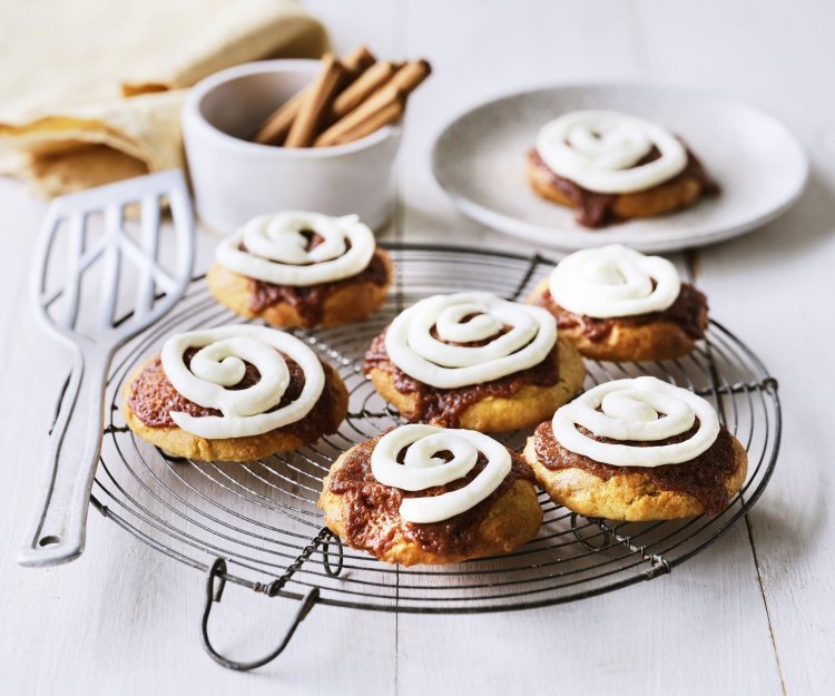 Op een taartrooster liggen 6 koekjes af te koelen. Het zijn koekjes van cinnamon rolls, een nieuwe versie om te genieten van deze heerlijke en kruidige zoetigheid. Op de koekjes is frosting gespoten op basis van roomkaas. 