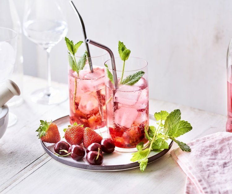 Twee cocktails staan op tafel, ze hebben een mooie roodroze kleur. Onderaan het glas ligt het smached fruit, met daarna de vodka en roze tonic. Er liggen nog enkele aardbeien en kersen langs. 