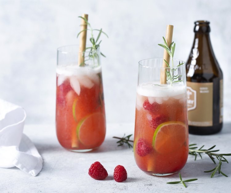 2 cocktails staan uitgeschonken in grote glazen. Deze cocktail op basis van bier en gin werd afgewerkt met frambozen en rozemarijn.