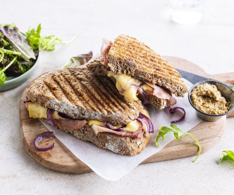 De croque ligt op een houten plankje aangesneden. De kaas en rode ui zien er heerlijk uit. In de achtergrond zie je ook nog wat gemengde sla en een potje graantjesmosterd. 