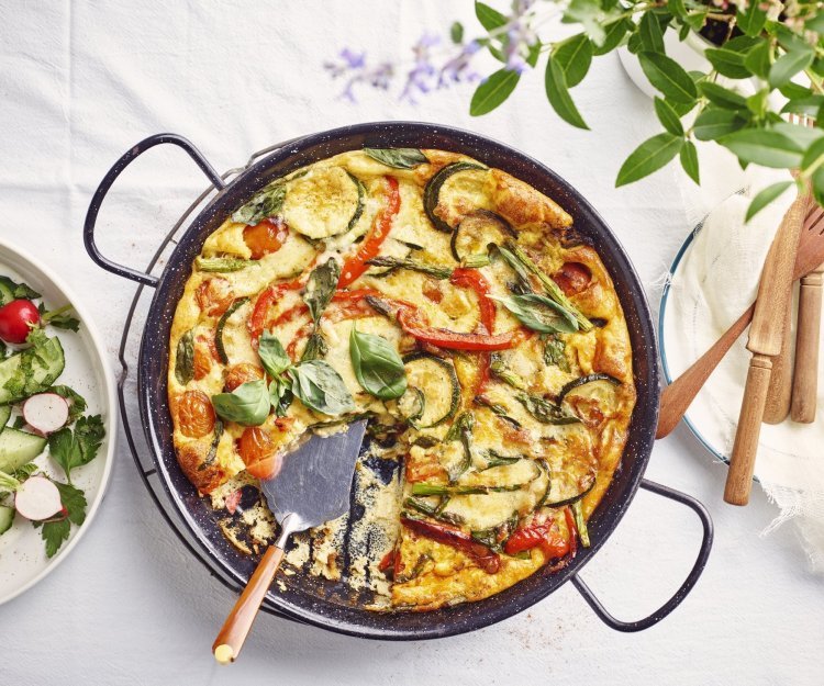 De frittata staat op tafel in zwarte pan, waardoor hij nog mooier oogt dan dat hij al is. De tomaatjes, paprika, courgette en asperges geven leuke kleurtjes aan de frittata. Er staat een slaatje langs de linkse zijde, met komkommer, radijsjes en verse kruiden.  