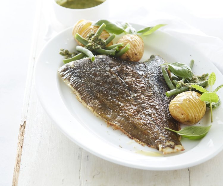 De gebakken pladijs op de graat ligt op een groot wit ovaal bord. Wij serveren hem met boontje langs en enkele hasselback potatoes. Het bord staat op een witte houten tafel en de salsa verde staat apart in een potje boven het bord.  