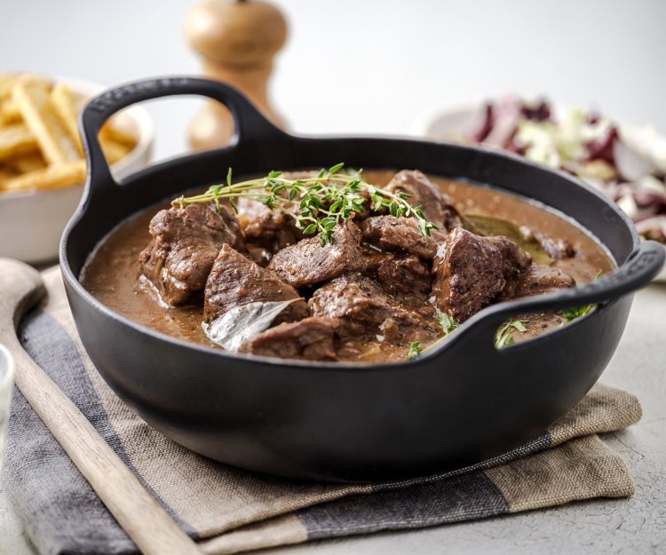 Er staat een zwarte sudderpot op tafel met Gentse stoverij, in de achtergrond zijn er frietjes te zien en een witloofslaatje.